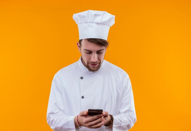 Un sonriente joven chef barbudo con uniforme blanco mirando su teléfono móvil mientras está de pie sobre una pared naranja