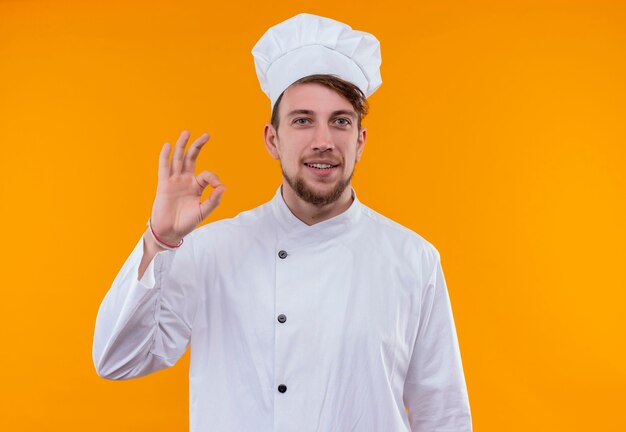 Un sonriente joven chef barbudo con uniforme blanco que muestra un gesto de ok con los dedos mientras mira en una pared naranja