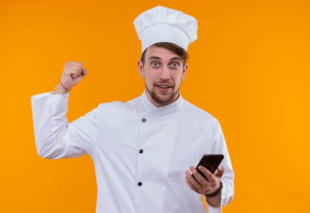 Un sonriente joven chef barbudo con uniforme blanco sosteniendo el teléfono móvil mientras mira con el puño cerrado en una pared naranja