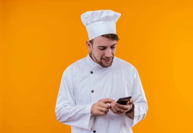 Un sonriente joven chef barbudo con uniforme blanco tocando su teléfono móvil en una pared naranja