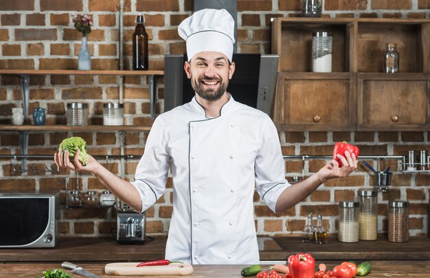 Sonriente joven chef masculino con brócoli y pimiento rojo en sus manos