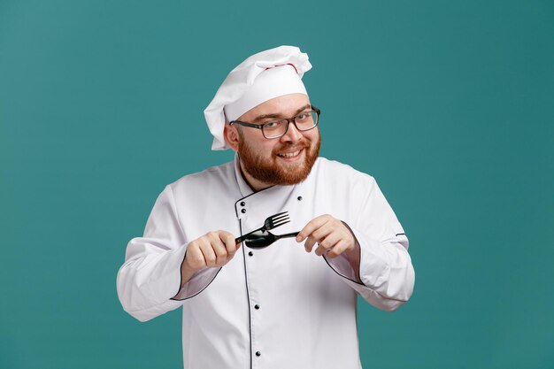 Sonriente joven chef masculino con uniforme de anteojos y gorra sosteniendo un tenedor y una cuchara mirando a la cámara frotándolos uno contra el otro aislado en el fondo azul