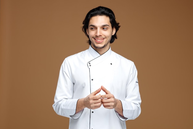 Sonriente joven chef masculino vistiendo uniforme manteniendo las manos juntas mirando al lado aislado sobre fondo marrón.