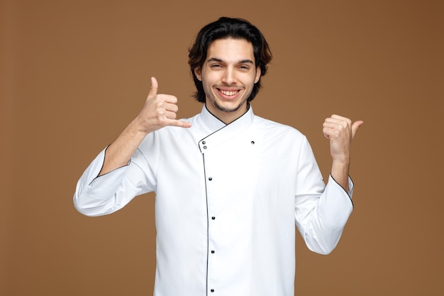 Sonriente joven chef uniformado mirando a la cámara mostrando gesto de llamada apuntando al lado aislado sobre fondo marrón.