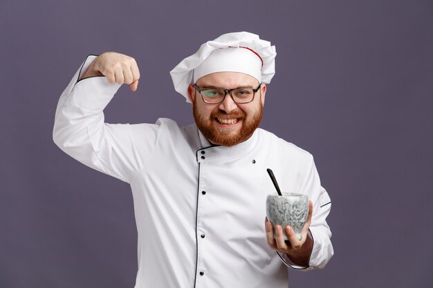Sonriente joven chef con uniforme de anteojos y gorra sosteniendo un tazón con una cuchara mirando a la cámara mostrando un gesto fuerte aislado en un fondo morado