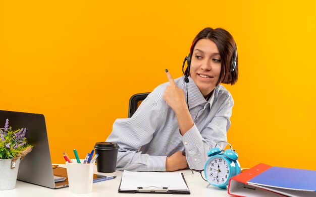 Sonriente joven chica del centro de llamadas con auriculares sentados en el escritorio mirando al lado apuntando detrás aislado en la pared naranja