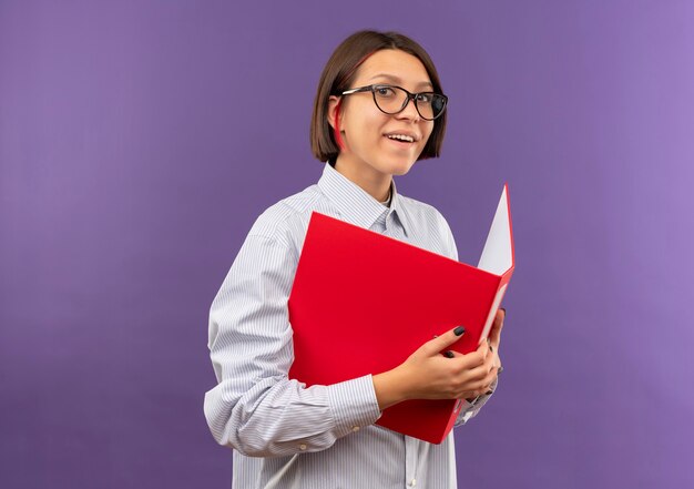 Sonriente joven chica de centro de llamadas con gafas sosteniendo la carpeta aislada en la pared púrpura