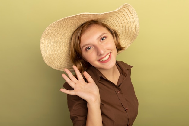 Sonriente joven chica rubia con sombrero de playa de pie en la vista de perfil mirando agitando aislado en la pared verde oliva con espacio de copia