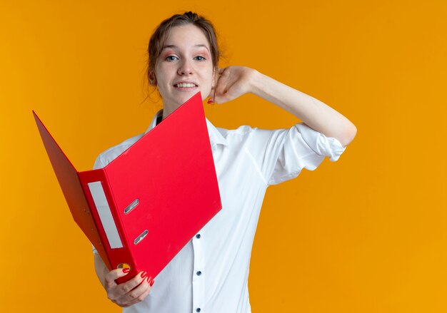 Sonriente joven chica rusa rubia pone la mano detrás de la cabeza sosteniendo la carpeta de archivos