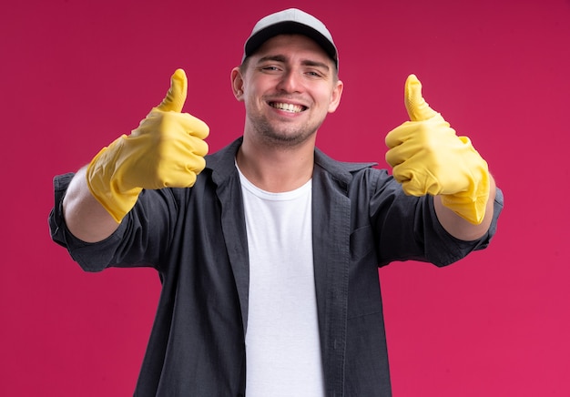 Sonriente joven chico de limpieza hamdsome con camiseta y gorra con guantes mostrando los pulgares para arriba aislado en la pared rosa