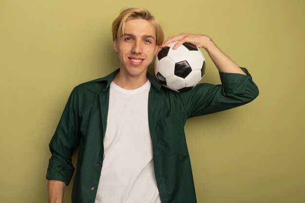 Sonriente joven chico rubio vistiendo camiseta verde poniendo la pelota en el hombro