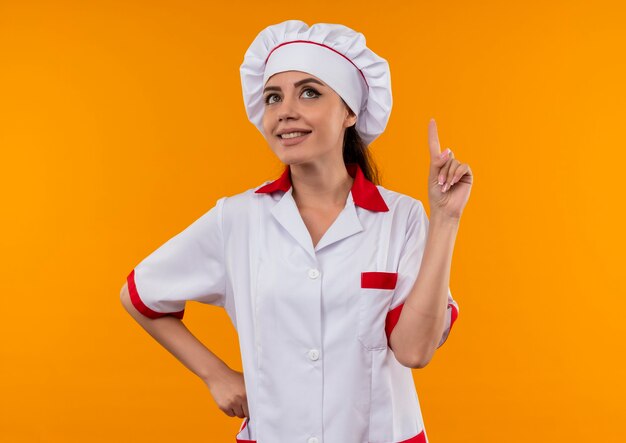 Sonriente joven cocinera caucásica en uniforme de chef apunta hacia arriba y pone la mano en la cintura aislada en la pared naranja con espacio de copia