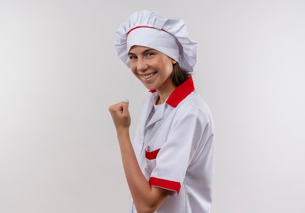 Sonriente joven cocinera caucásica en uniforme de chef se coloca de lado y mantiene el puño en blanco con espacio de copia