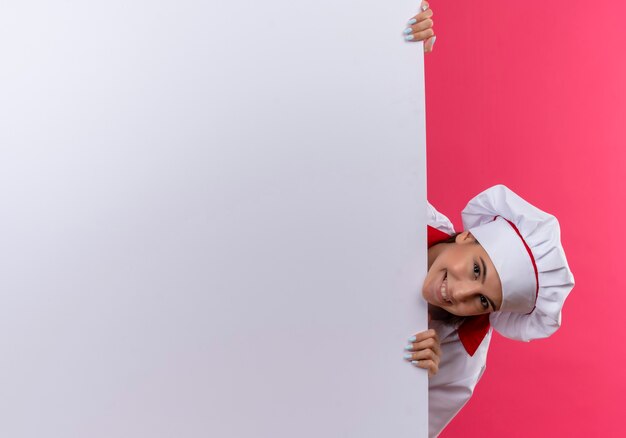 Sonriente joven cocinera caucásica en uniforme de chef está detrás de la pared blanca en rosa con espacio de copia