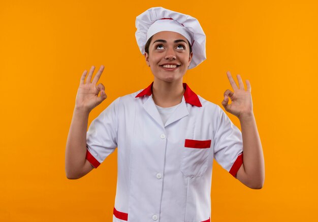 Sonriente joven cocinera caucásica en uniforme de chef gestos ok signo de mano y mirar hacia arriba aislado en la pared naranja con espacio de copia