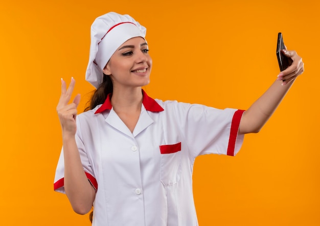 Sonriente joven cocinera caucásica en uniforme de chef mira teléfono y gestos signo de mano de victoria aislado en pared naranja con espacio de copia