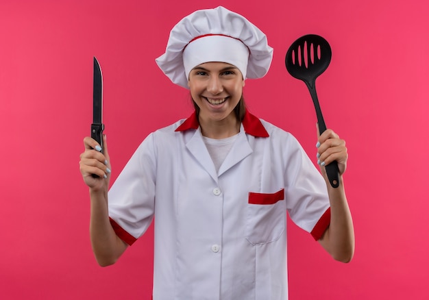 Sonriente joven cocinera caucásica en uniforme de chef sostiene cuchillo y espátula aislado en espacio rosa con espacio de copia