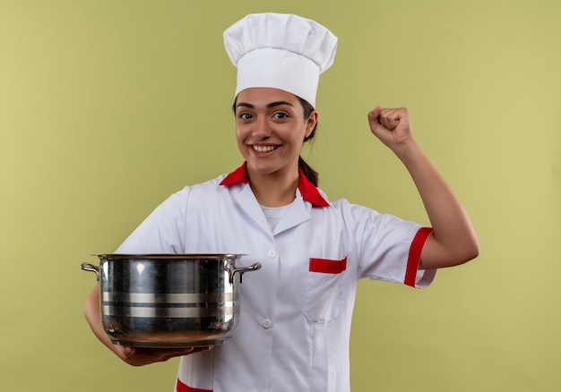 Sonriente joven cocinera caucásica en uniforme de chef sostiene la olla y levanta el puño aislado en la pared verde con espacio de copia