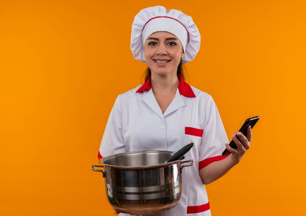 Sonriente joven cocinera caucásica en uniforme de chef sostiene olla y teléfono aislado en pared naranja con espacio de copia