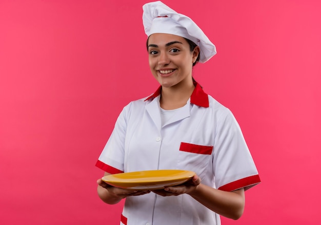 Sonriente joven cocinera caucásica en uniforme de chef sostiene el plato con ambas manos aislado en la pared rosa con espacio de copia