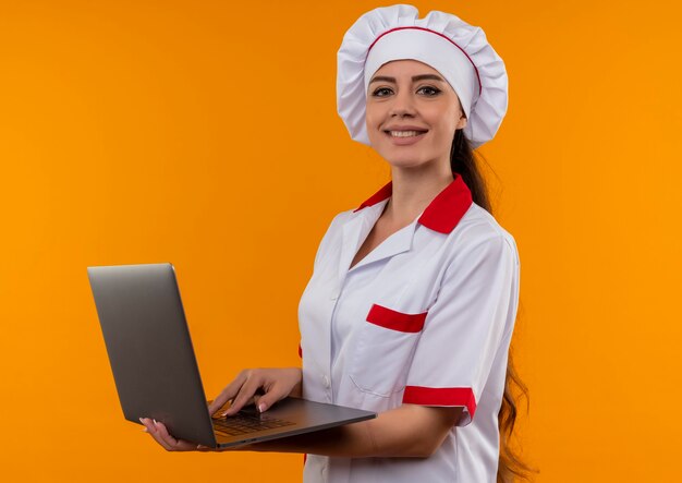 Sonriente joven cocinera caucásica en uniforme de chef sostiene portátil y mira a cámara aislada en pared naranja con espacio de copia