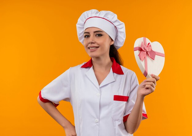 Sonriente joven cocinera caucásica en uniforme de chef tiene caja en forma de corazón aislada en pared naranja con espacio de copia