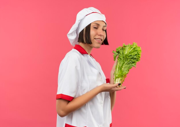 Sonriente joven cocinera en uniforme de chef de pie en la vista de perfil sosteniendo y apuntando con lechuga de mano aislado en la pared rosa