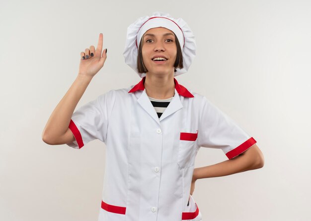 Sonriente joven cocinera en uniforme de chef poniendo la mano en la cintura y levantando el dedo aislado en la pared blanca