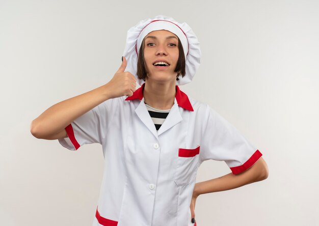 Sonriente joven cocinera en uniforme de chef poniendo la mano en la cintura y mostrando el pulgar hacia arriba aislado en la pared blanca