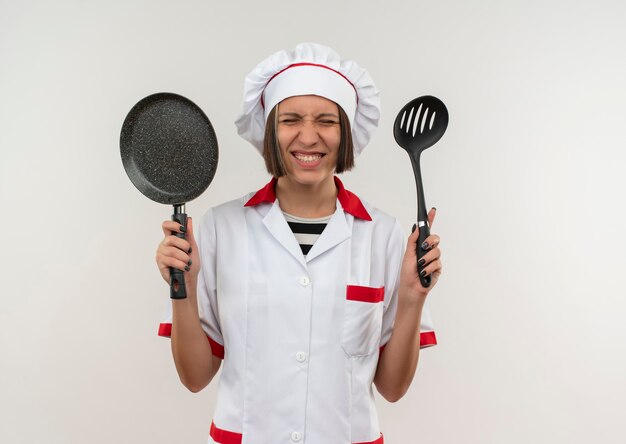 Sonriente joven cocinera en uniforme de chef sosteniendo una espátula y una sartén con los ojos cerrados aislados en la pared blanca