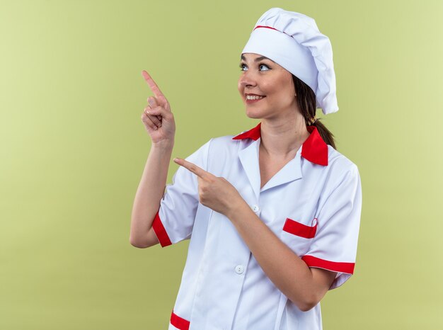 Sonriente joven cocinera vistiendo puntos de uniforme de chef en el lado aislado en la pared verde oliva con espacio de copia