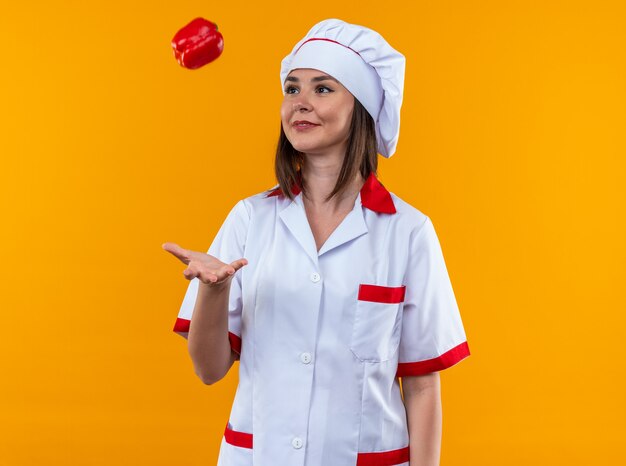 Sonriente joven cocinera vistiendo uniforme de chef arrojando pimienta aislado en la pared naranja