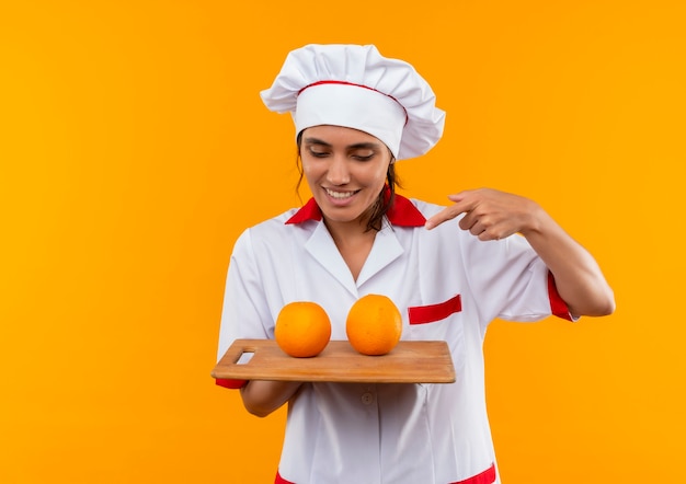Sonriente joven cocinera vistiendo uniforme de chef sosteniendo y apunta a naranja en la tabla de cortar con espacio de copia