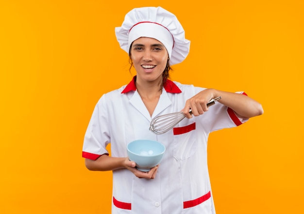 Sonriente joven cocinera vistiendo uniforme de chef sosteniendo un batidor y un tazón con espacio de copia
