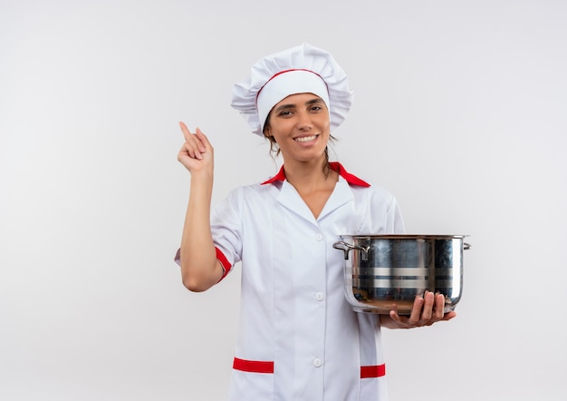 Sonriente joven cocinera vistiendo uniforme de chef sosteniendo una cacerola y apunta al lado con espacio de copia