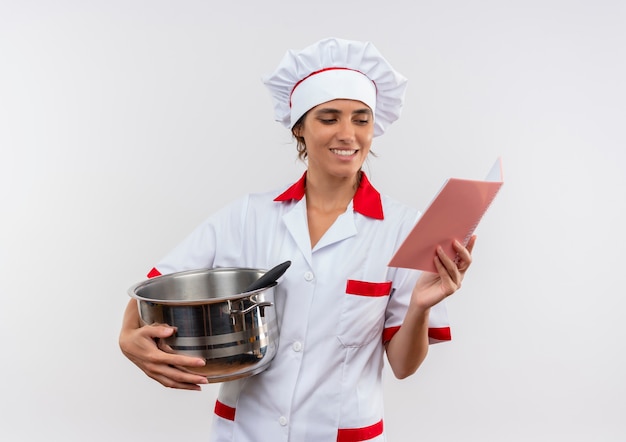 Sonriente joven cocinera vistiendo uniforme de chef sosteniendo una cacerola y lookingc en el cuaderno en la mano con espacio de copia