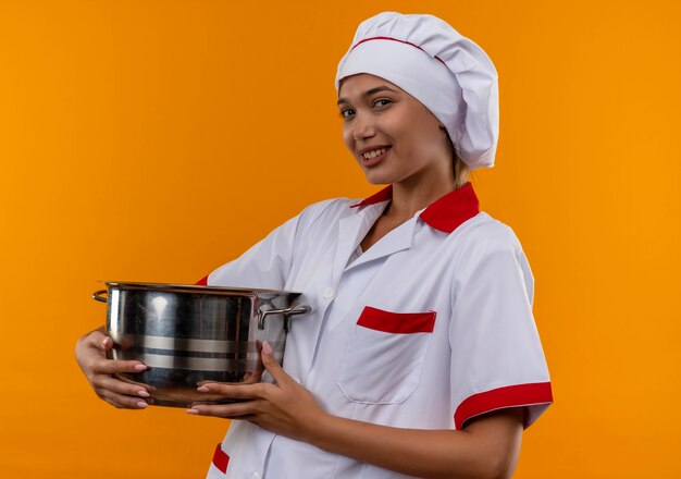 Sonriente joven cocinera vistiendo uniforme de chef sosteniendo una cacerola sobre fondo naranja aislado