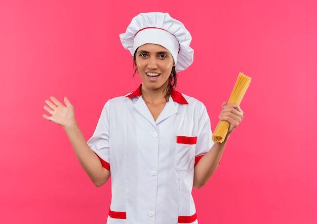 Sonriente joven cocinera vistiendo uniforme de chef sosteniendo espaguetis y extendiendo la mano con espacio de copia