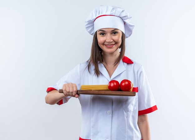 Sonriente joven cocinera vistiendo uniforme de chef sosteniendo espaguetis con tomates en la tabla de cortar aislado en la pared blanca