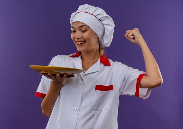 Sonriente joven cocinera vistiendo uniforme de chef sosteniendo la placa mostrando sí gesto sobre fondo aislado con espacio de copia