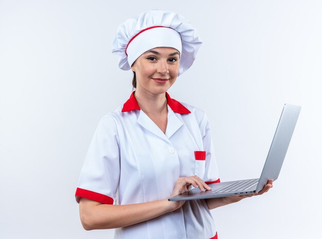 Sonriente joven cocinera vistiendo uniforme de chef sosteniendo portátil aislado en la pared blanca