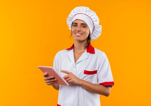 Sonriente joven cocinera vistiendo uniforme de chef sosteniendo y puntos cuaderno con espacio de copia