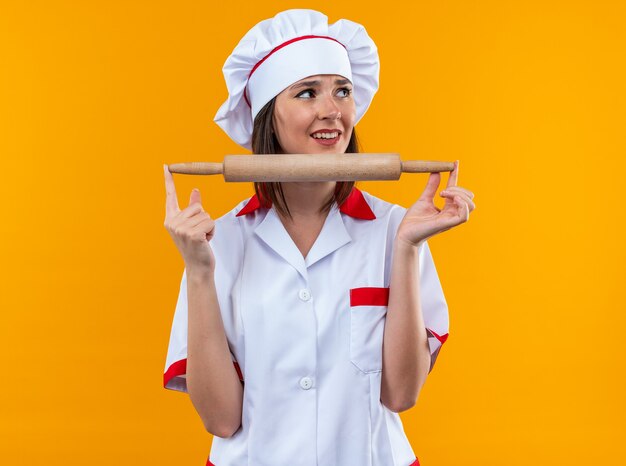 Sonriente joven cocinera vistiendo uniforme de chef sosteniendo rodillo aislado sobre fondo naranja