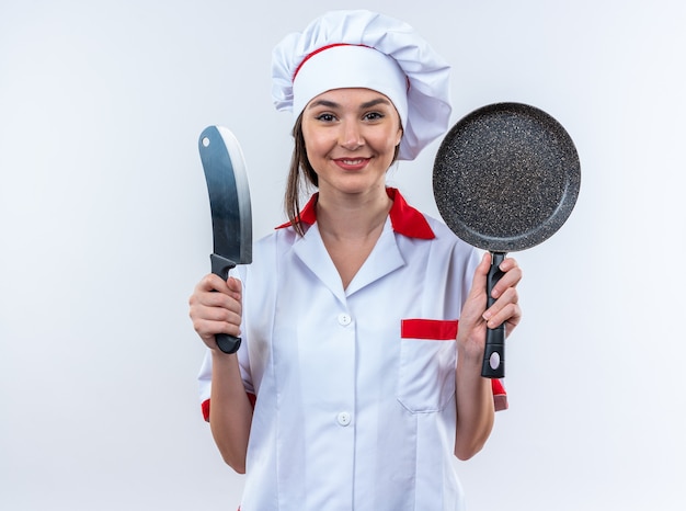 Sonriente joven cocinera vistiendo uniforme de chef sosteniendo una sartén con cuchilla aislado sobre fondo blanco.