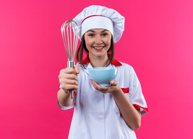 Sonriente joven cocinera vistiendo uniforme de chef sosteniendo el tazón con batidor en la cámara aislada sobre fondo de color rosa