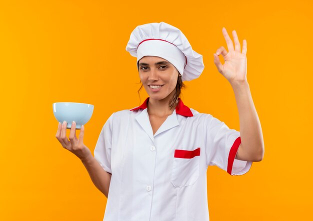 Sonriente joven cocinera vistiendo uniforme de chef sosteniendo el tazón y mostrando gesto okey