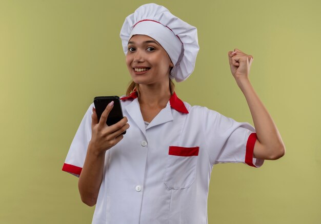 Sonriente joven cocinera vistiendo uniforme de chef sosteniendo el teléfono mostrando sí gesto