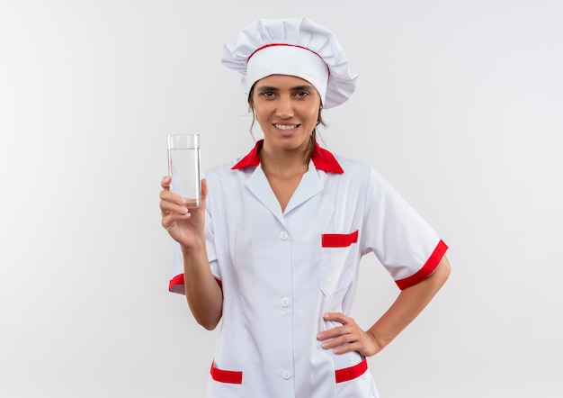 Sonriente joven cocinera vistiendo uniforme de chef sosteniendo un vaso de agua poniendo la mano en la cadera con espacio de copia