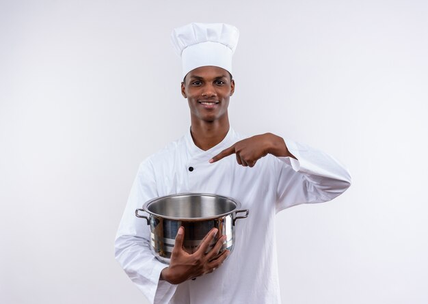 Sonriente joven cocinero afroamericano en uniforme de chef sostiene la cacerola y apunta a la cacerola sobre fondo blanco aislado con espacio de copia