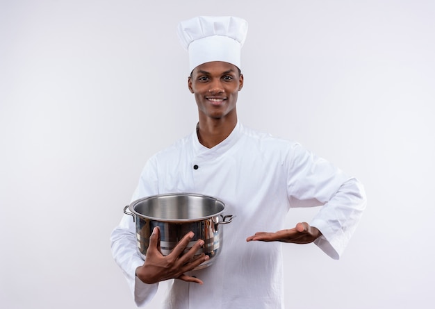 Sonriente joven cocinero afroamericano en uniforme de chef sostiene una cacerola y mantiene la mano recta sobre fondo blanco aislado con espacio de copia